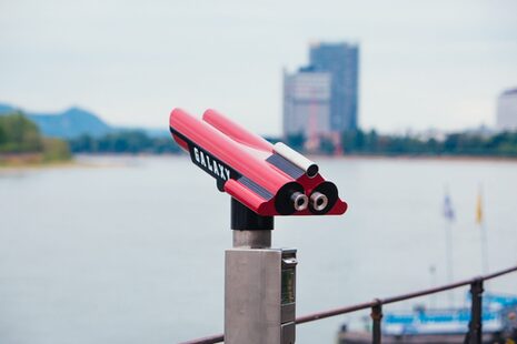 Ein rotes Fernrohr am Rheinufer, im Hintergrund der UN Campus