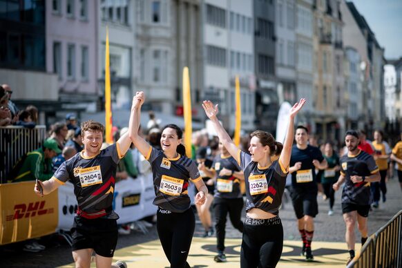 The photo shows three young people in sportswear raising their arms in the air with joy. They are at the finish line