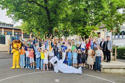 Eine große Gruppe winkender Kinder steht mit einigen Erwachsenen und einer als Löwe verkleideten Person unter einem großen Baum auf einem Schulhof. Eine als Eisbär verkleidete Person liegt quer vor der Gruppe auf dem Boden und winkt ebenfalls.gelben
