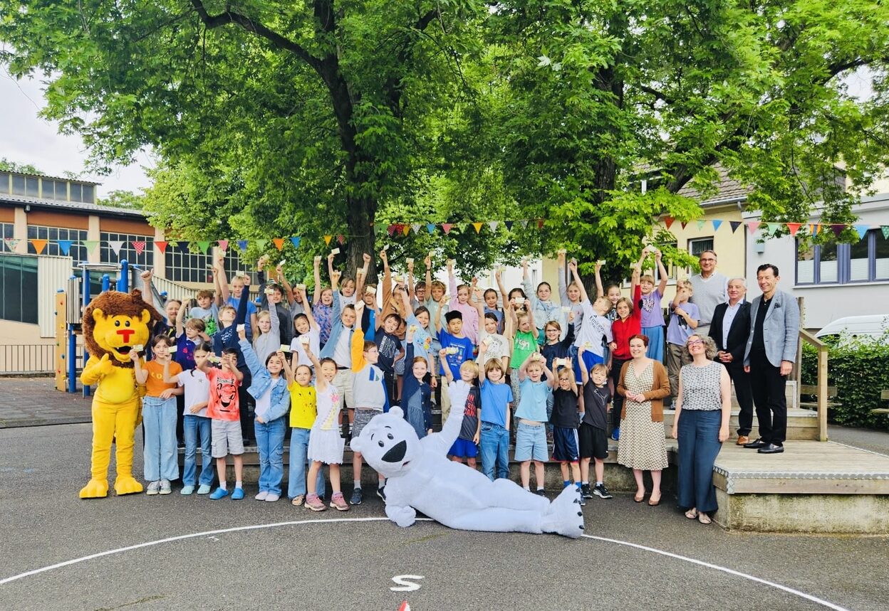 Eine große Gruppe winkender Kinder steht mit einigen Erwachsenen und einer als Löwe verkleideten Person unter einem großen Baum auf einem Schulhof. Eine als Eisbär verkleidete Person liegt quer vor der Gruppe auf dem Boden und winkt ebenfalls.