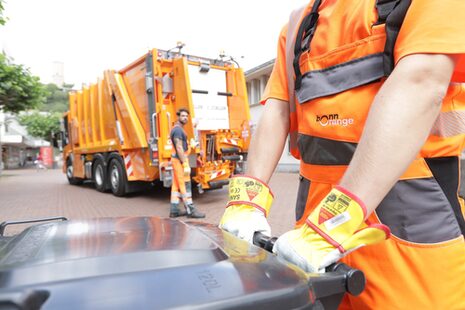 Abfallsammelfahrzeug der bonnorange AöR in der Fußgängerzone Bad Godesberg mit einer Restabfalltonne, die ein Mitarbeiter von bonnorange mit beiden Händen in Arbeitshandschuhen zu sich zieht.