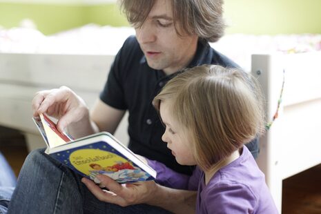 Ein Mann sitzt neben einem Mädchen auf dem Boden und liest aus einem Buch vor