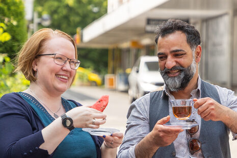 Zwei Personen sitzen im Freien an einem Tisch. Sie trinken Tee und essen Wassermelone.