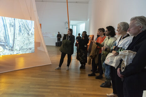 Besucher:innen einer Videonale-Ausstellung stehen vor einer Projektion, während eine Führung stattfindet.