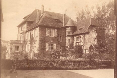 تُظهر الصورة قلعة ليدي في حوالي عام 1900 في صورة فوتوغرافية قديمة بلون بني داكن. هناك هندسة معمارية ببرج موضوع بين جانبي المبنى، ونافذة كبيرة مع شرفة فوقها، وبوابة القلعة ومصاريع.