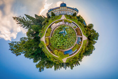 Das Fotozeigt in einer Fischaugen-Rundumaufnahme das Poppelsdorfer Schloss mit Teilen des Parks vor blauem Himmel. Der Eindruck entsteht, als handelt es sich um eine Kugel, die im Himmel schwebt, ähnlich der Erde im All.